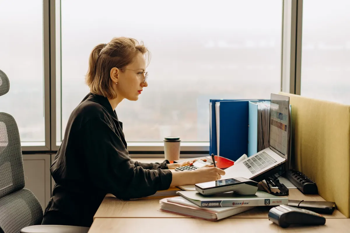 An accountant at a desk by a window, with binders, a calculator and a laptop
