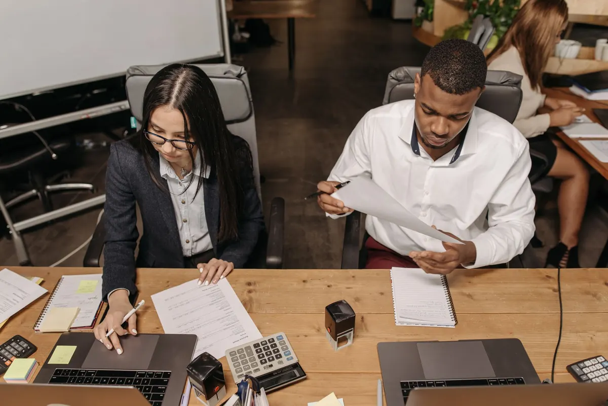Two accountants at a wooden desk with documents, calculators and stamps